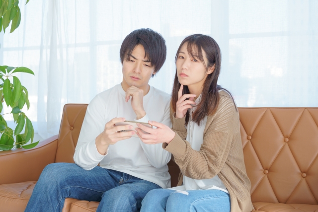 ソファでスマートフォンを見ながら考え込む夫婦